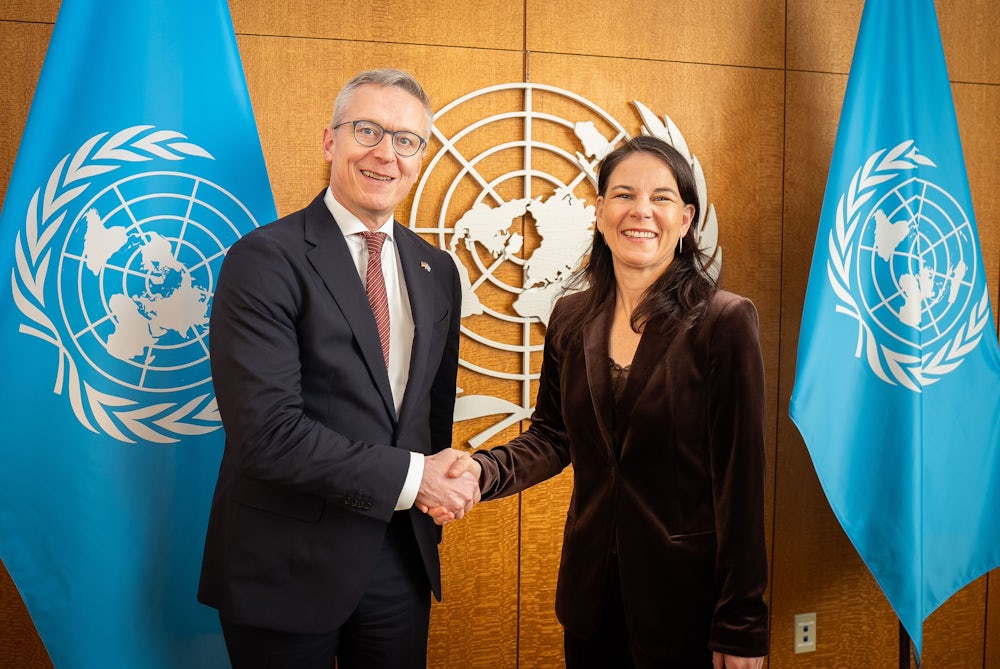 Karsten Wildberger shaking hands with a woman in a dark velvet jacket in front of a wood-panelled wall bearing the United Nations emblem, flanked by two light blue UN flags