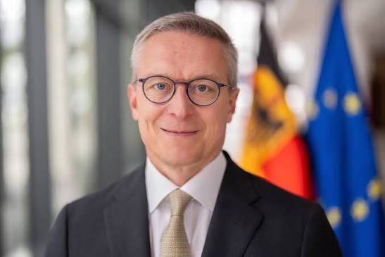 Official portrait of Karsten Wildberger in a dark suit and beige tie, smiling, with the German and European flags behind him