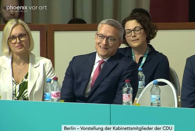 Karsten Wildberger smiling while seated in a row of people at a podium table, in a television still captioned Berlin – Vorstellung der Kabinettsmitglieder der CDU