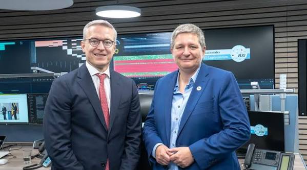 Karsten Wildberger standing beside a colleague in front of large monitoring screens in a control room bearing the logo of the Federal Office for Information Security, BSI