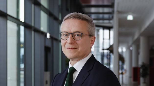 Karsten Wildberger in a dark suit and green tie, wearing glasses, standing in a bright modern corridor with glass walls