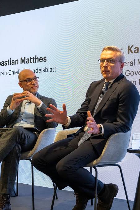 Karsten Wildberger seated on stage in a grey armchair, gesturing as he speaks, next to the Handelsblatt editor-in-chief Sebastian Matthes, whose name appears on the screen behind them