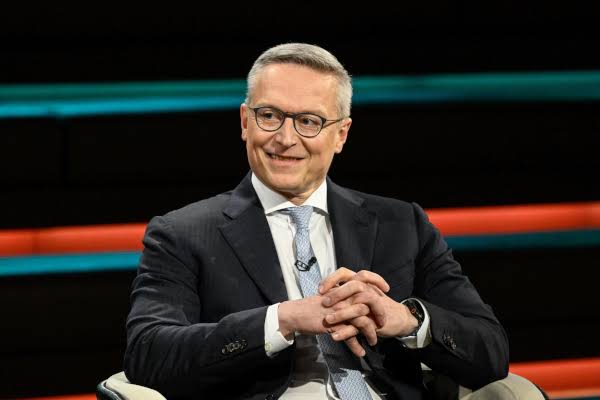 Karsten Wildberger seated in a television studio, smiling with hands clasped, in a dark suit and light blue tie, with horizontal coloured light bands behind him
