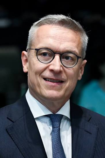 Portrait of Karsten Wildberger in a dark suit and patterned blue tie, wearing round glasses and smiling