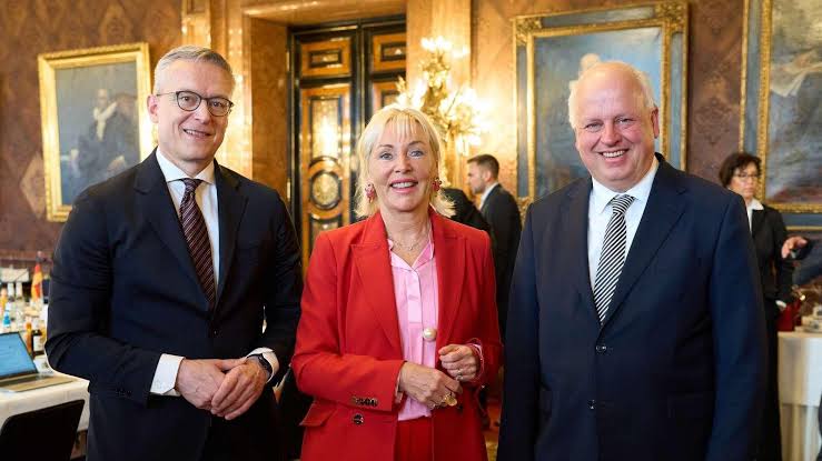 Karsten Wildberger standing with a woman in a red jacket and a man in a dark suit in an ornate historic hall with large paintings and a chandelier