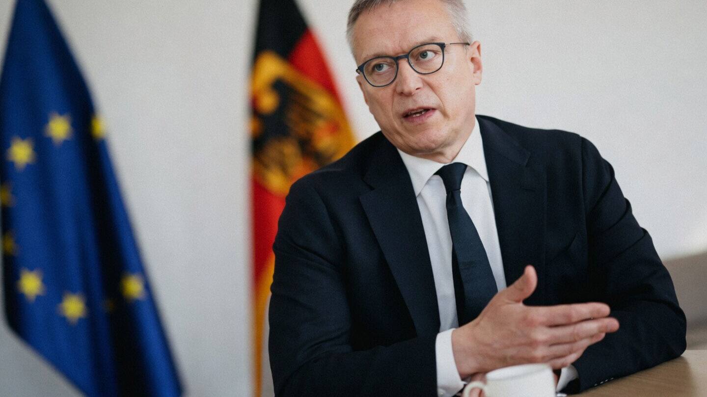 Karsten Wildberger seated at a table during an interview, gesturing with one hand, with the European and German flags in the background