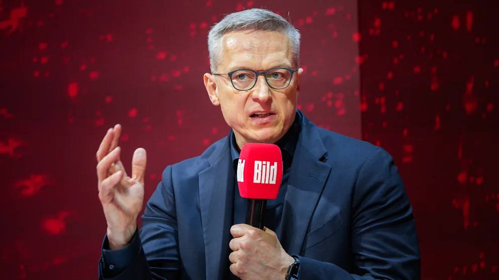 Karsten Wildberger speaking on stage, holding a red microphone with the BILD logo and gesturing with his other hand, against a dark red background
