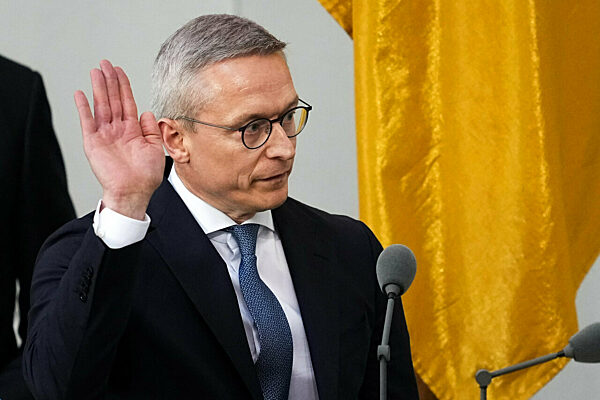 Karsten Wildberger raising his right hand to take the oath of office in the Bundestag, in front of a gold-coloured flag and microphones
